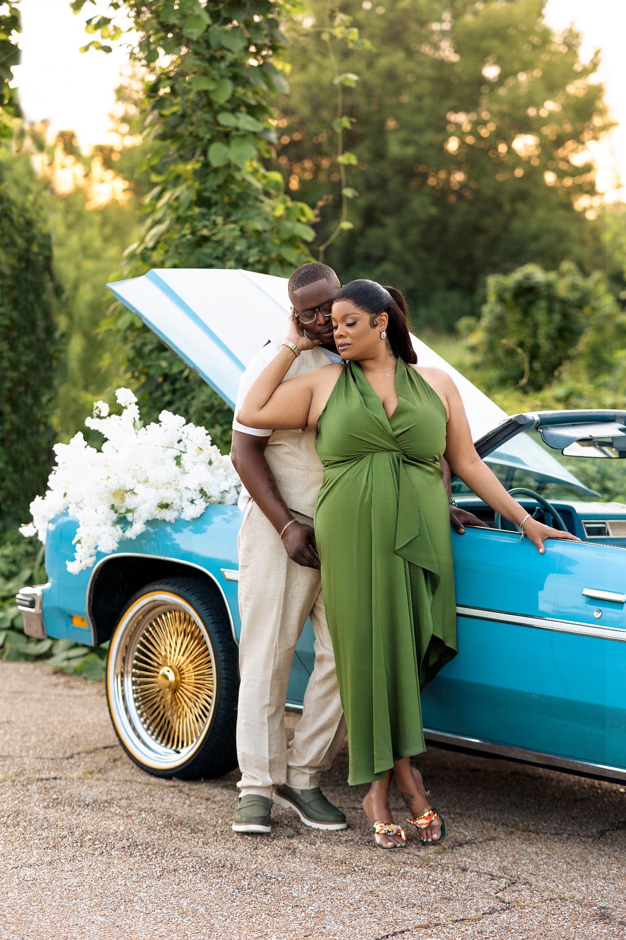 Engaged couple embracing beside a classic blue convertible at sunset